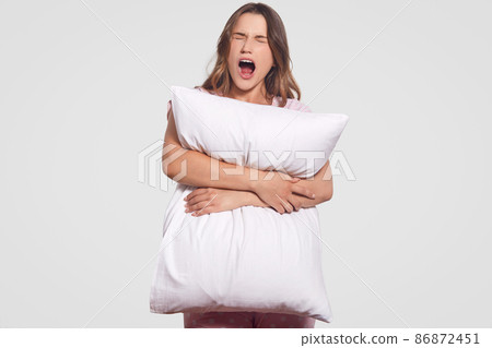 Annoyed desperate young woman yells loudly, keeps mouth widely opened, holds white pillow, poses against studio background, dressed in pyjamas. People, negative emotions and feelings concept Annoyed desperate young woman yells loudly, keeps mouth widely opened, holds white pillow, poses against studio background, dressed in pyjamas. People, negative emotions and feelings concept 86872451