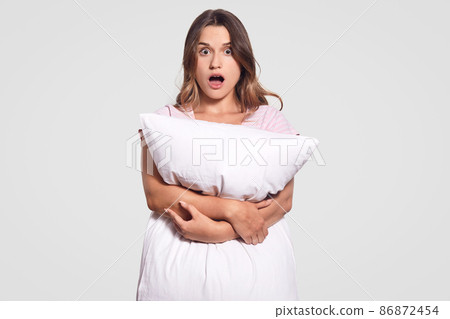 Astonished dark haired woman carries white soft pillow, horrified by nightmare, surprised by something terrible, keeps jaw dropped, looks with fear, isolated over white background. Sleeping concept Astonished dark haired woman carries white soft pillow, horrified by nightmare, surprised by something terrible, keeps jaw dropped, looks with fear, isolated over white background. Sleeping concept 86872454