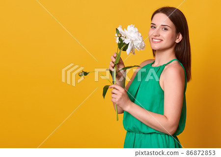 Studio shot of charming woman wearing elegant green sundress isolated over yellow studio background, holding white peony flower, having pleased expression. Copy space for advertisment or promotion. 86872853