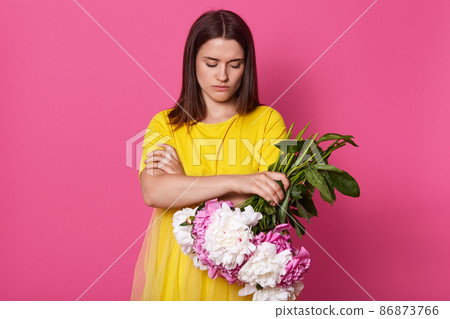 Indoor shot of beautiful brunette woman holding flowers isolated over pink studio background,lady wearing yellow dress, darkhaired female with folded hands, looks offended, quarrels with boyfriend. Indoor shot of beautiful brunette woman holding flowers isolated over pink studio background,lady wearing yellow dress, darkhaired female with folded hands, looks offended, quarrels with boyfriend. 86873766