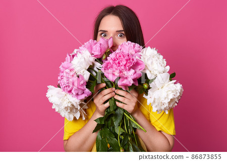 Portrait of energetic attractive lady with dark hair standing isolated over pink background, pressing flowers close to body, covering almost all part of face with peonies, opening eyes widely. 86873855