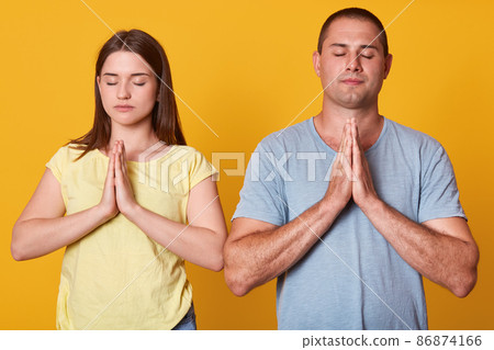Indoor shot of peaceful beautiful couple standing isolated over yellow background in studio, closing their eyes, putting their hands together, making gesture, wearing casual items of clothes. Indoor shot of peaceful beautiful couple standing isolated over yellow background in studio, closing their eyes, putting their hands together, making gesture, wearing casual items of clothes. 86874166