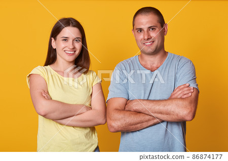 Handsome cheerful man standing near his girlfriend with folded arms, smiling sincerely, being in high spirits, being on good terms, looking directly at camera, wearing casual t shirts. Couple concept. 86874177