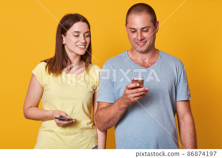 Handsome man with mobile phone in hand, checking his social network, stands near cute playful brunette woman which looking in his smart phone. Positive couple isolated over yellow background. Handsome man with mobile phone in hand, checking his social network, stands near cute playful brunette woman which looking in his smart phone. Positive couple isolated over yellow background. 86874197