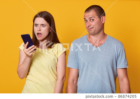 Image of astonished woman holding husband's phone, checking his messages with open mouth and shocked facial expression, man looks at camera with perplexity, isolated over yellow studio background. 86874205