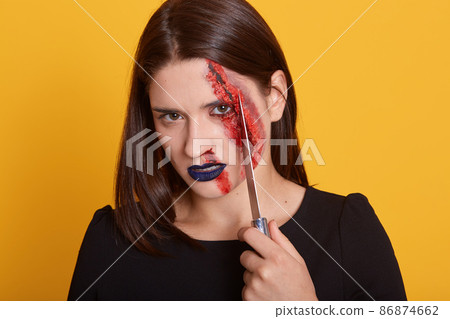 Indoor shotof of girl all in blood, lady holding knife near her visage, angry woman having gloomy makeup, beautiful woman has dangerous facial expression, wearing blackoutfit. Halloween concept. Indoor shotof of girl all in blood, lady holding knife near her visage, angry woman having gloomy makeup, beautiful woman has dangerous facial expression, wearing blackoutfit. Halloween concept. 86874662