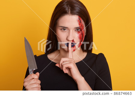 Horizontal shot of young stylish brunette girl with halloween bloody makeup, standing and looking straight with finger near her mouth, showssilent, posing over yellow background. Halloween concept. Horizontal shot of young stylish brunette girl with halloween bloody makeup, standing and looking straight with finger near her mouth, showssilent, posing over yellow background. Halloween concept. 86874663