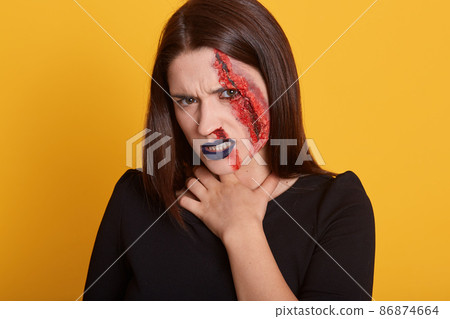Indoor shot of young brunette girl with stylish dark halooween make up, with blood that flows from her temple and cheek, female wearing black t shirt, keeps hand on her neck, halloween concept. Indoor shot of young brunette girl with stylish dark halooween make up, with blood that flows from her temple and cheek, female wearing black t shirt, keeps hand on her neck, halloween concept. 86874664