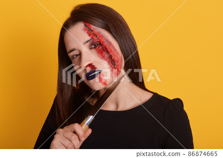 Indoor shot of brunette female with knife near her neck and bloody wound on her face, looking directly at camera, dresses black shirt, posing isolated over yellow studio background. Halloween concept. Indoor shot of brunette female with knife near her neck and bloody wound on her face, looking directly at camera, dresses black shirt, posing isolated over yellow studio background. Halloween concept. 86874665