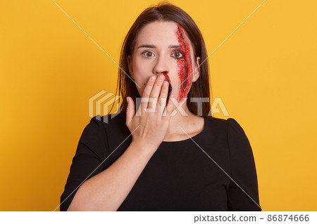Indoor shot of beautiful young woman covering her mouth with hand, isolated over yellow background, has bloody wound on her cheek, looks shocked and astonished, dresses black shirt. Halloween concept. Indoor shot of beautiful young woman covering her mouth with hand, isolated over yellow background, has bloody wound on her cheek, looks shocked and astonished, dresses black shirt. Halloween concept. 86874666