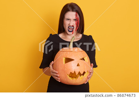 Image of serious young woman wearing black dress, looking screaming directly at camera, lady expresses anger, girl in halloween costume isolated over yellow background with pumpkin in hands. 86874673