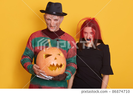 Portrait of man and woman with halloween pumpkin against yellow studio background, male with knives instead of fingers and female in black dress and red veil, look at camera with angry dangerous look. 86874710
