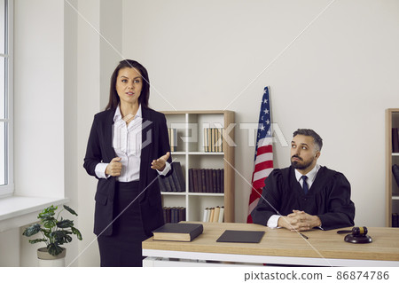 Lawyer or defense attorney making speech during trial session in court. Serious unsmiling woman who represents defendant in lawsuit or criminal prosecution speaking to judge and audience in courtroom Lawyer or defense attorney making speech during trial session in court. Serious unsmiling woman who represents defendant in lawsuit or criminal prosecution speaking to judge and audience in courtroom 86874786