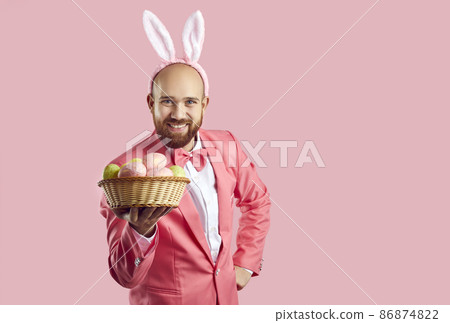 Portrait of happy funny handsome bearded young man in pink suit, bow tie and cute bunny ears standing isolated on color background, looking at camera, showing wicker bowl with Easter eggs and smiling Portrait of happy funny handsome bearded young man in pink suit, bow tie and cute bunny ears standing isolated on color background, looking at camera, showing wicker bowl with Easter eggs and smiling 86874822
