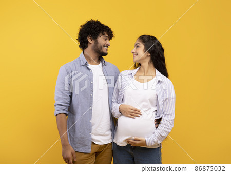 Happy indian couple expecting baby, posing over yellow background, embracing and looking at each other Happy indian couple expecting baby, posing over yellow background, embracing and looking at each other 86875032