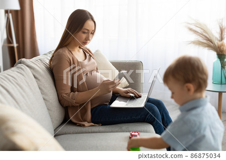 Young pregnant freelancer woman using smartphone and laptop, mom taking care of her toddler son Young pregnant freelancer woman using smartphone and laptop, mom taking care of her toddler son 86875034