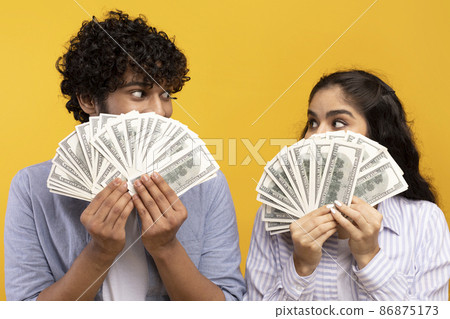 Big win concept. Young indian couple holding and covering faces with a lot of dollar cash fan, yellow studio background 86875173