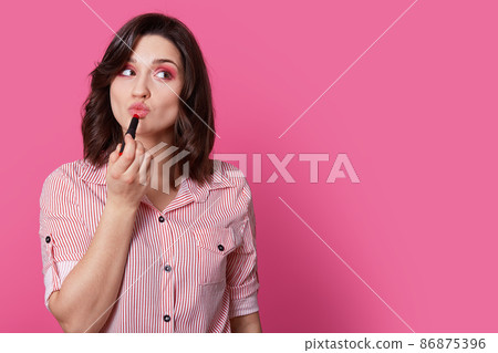 Image of beautiful young woman with makeup, keeps lips folded, uses lipstick, cares of her beauty, wears stylish shirt, isolated over pink background with copy space for your promotional content Image of beautiful young woman with makeup, keeps lips folded, uses lipstick, cares of her beauty, wears stylish shirt, isolated over pink background with copy space for your promotional content 86875396