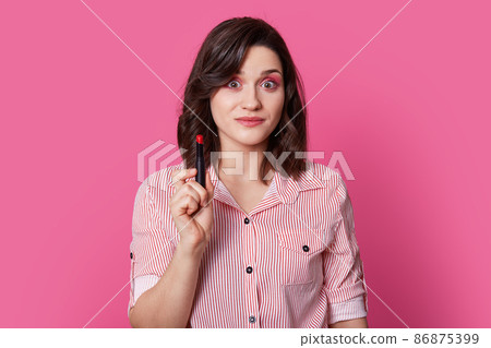Photo of surprised brunette young woman with trendy hairstyle, has shocked expression, holds red lipstick, wears striped shirt, isolated over bright pink background. People and beauty concept Photo of surprised brunette young woman with trendy hairstyle, has shocked expression, holds red lipstick, wears striped shirt, isolated over bright pink background. People and beauty concept 86875399