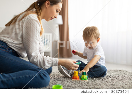 Young mom and toddler child playing together at home, using toys while sitting on floor carpet in living room 86875404