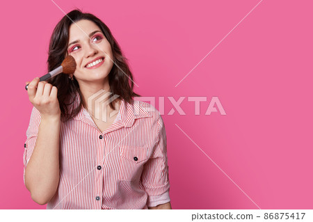 Isolated shot of pleasant looking young woman with cheerful expression, keeps cosmetic brush on cheek, looks dreamfully upwards, thinks about something pleasant, isolated over rosy studio wall. 86875417