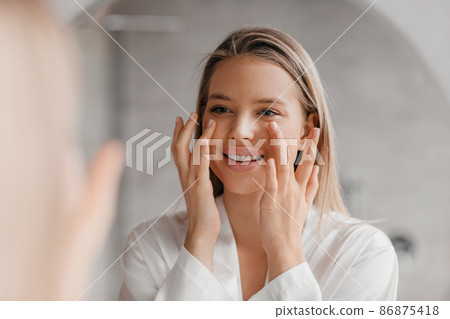Pretty lady making face-lifting massage in bathroom, looking at mirror and smiling, using moisturizing face cream 86875418