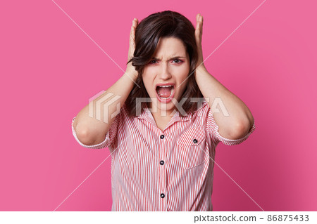 Frustrated brunette lady yells with anger, covers ears, annoyed with loud noise, wears striped shirt, feels outraged, dressed in striped clothes, isolated over bright pink background. Stop this sound 86875433