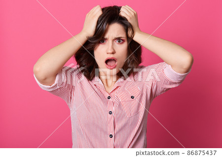 Frustrated confused Caucasian woman keeping hands in her hair, posing with widely opened mouth, wearing striped shirt, being in bad mood, looks shocked. Frustrated confused Caucasian woman keeping hands in her hair, posing with widely opened mouth, wearing striped shirt, being in bad mood, looks shocked. 86875437