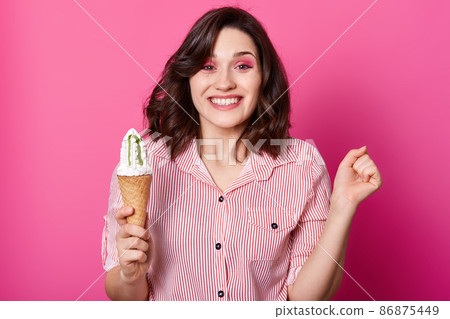 Studio shot of cheerful dark haired woman holds ice cream, clench fist, has happy facial expression, posing in striped shirt, has toothy smile, isolated on rose background. Happyness, emotions concept 86875449