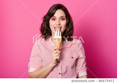 Image of happy woman eating ice cream, has dark wavy hair, wearing stylish shirt with red stripes, has bright makeup, looking at camera with happy expression, isolated on rose studiobackground. 86875451