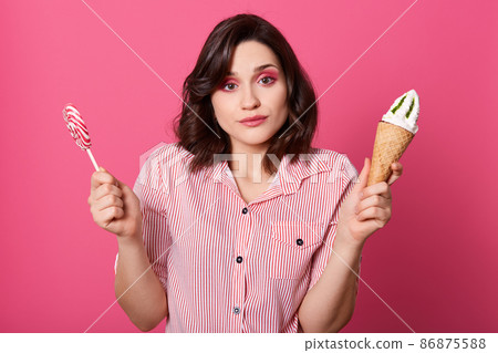 Photo of confused pensive sweet female holding ice cream and lollipop in both hands, looking directly at camera, having bright makeup, making choice, having thoughtful facial expression. Food concept. 86875588