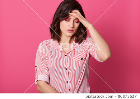 Close up portrait of dissatisfied brunette woman touches her forehead with palm, posing with closed eyes, suffers from migraine, wears striped blouse, young female closes eyes from bad feelings. Close up portrait of dissatisfied brunette woman touches her forehead with palm, posing with closed eyes, suffers from migraine, wears striped blouse, young female closes eyes from bad feelings. 86875627