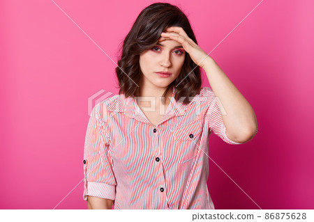 Image of young woman with terrible headache, keeps hand on forehead, needs to take medicine, studio photo of brunette girl in striped blouse, isolated over rosy background. People and health concept. Image of young woman with terrible headache, keeps hand on forehead, needs to take medicine, studio photo of brunette girl in striped blouse, isolated over rosy background. People and health concept. 86875628