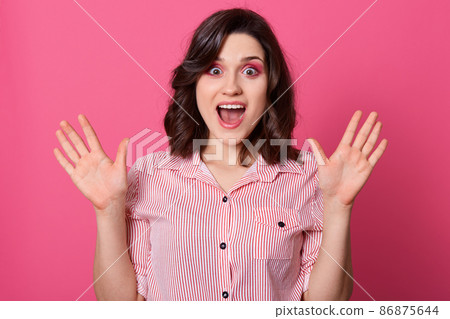 Horizontal shot of beautiful caucasian woman with shocked facial expression, keeps hands up, having widely opened eyes and mouth, has dark hair, wearing casual shirt, posing against pink wall. 86875644