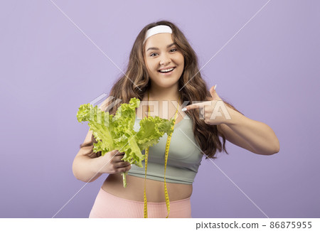 Portrait of happy young european plus size woman in sportswear with measuring tape points finger at salad Portrait of happy young european plus size woman in sportswear with measuring tape points finger at salad 86875955
