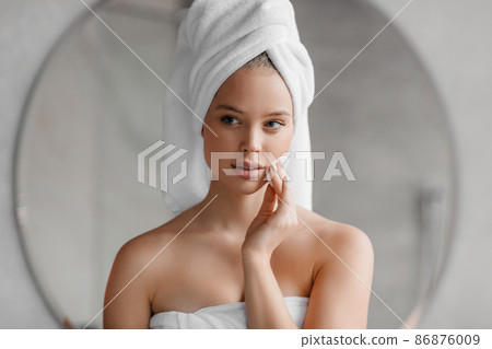 Beauty routine. Pretty young woman cleaning her face with cotton pad at home, standing with towel on head after bath 86876009