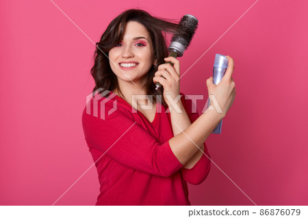 Good looking cheerful female styling her hair with hairbrush and spray, tangling hair with comb, standing isolated over pink background in studio, smiling sincerely. People and beauty concept. Good looking cheerful female styling her hair with hairbrush and spray, tangling hair with comb, standing isolated over pink background in studio, smiling sincerely. People and beauty concept. 86876079