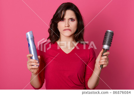 Image of desperate beautiful woman posing with pouty lips, cant make hairstyle, being upset and sad, carries comb and spray, has dark hair, models posing over pink studio wall, looks at camera. 86876084