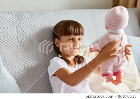 Dark haired female child wearing white t shirt, has pigtails raising her doll, looking at her toy with smile, playing alone while sitting on sofa at home. Dark haired female child wearing white t shirt, has pigtails raising her doll, looking at her toy with smile, playing alone while sitting on sofa at home. 86876515