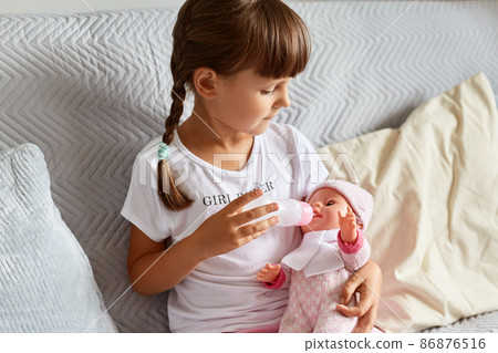 Cute kid at home sitting on sofa. Little girl with pigtails in nursery school, preschooler female child playing with doll, feeding her tiny daughter with bottle like mother. 86876516