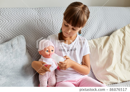 Indoor shot of little girl sitting on the sofa in the room at home playing with baby doll, like mom, dark haired preschooler female child holding toy. Indoor shot of little girl sitting on the sofa in the room at home playing with baby doll, like mom, dark haired preschooler female child holding toy. 86876517
