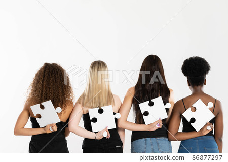 Four young girls of mixed ethnicity hold puzzles in their hands behind their backs with their backs to the camera. 86877297