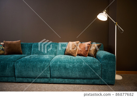 Exhibition of modern stylish upholstered furniture in the showroom of a furniture store. Focus on a turquoise soft velour sofa and brown pillows lit by a lamp against a brown wall background Exhibition of modern stylish upholstered furniture in the showroom of a furniture store. Focus on a turquoise soft velour sofa and brown pillows lit by a lamp against a brown wall background 86877682