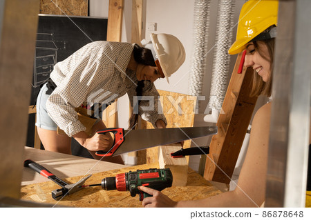 A brunette cuts a wooden log with a hand saw. Smiling girl while working on a construction site. A brunette cuts a wooden log with a hand saw. Smiling girl while working on a construction site. 86878648