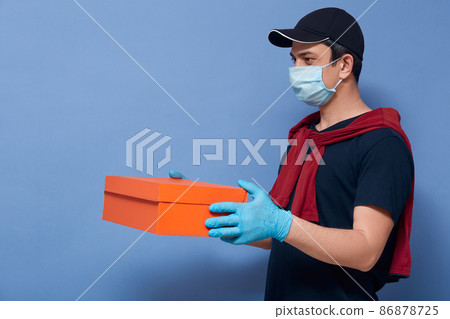 Profile of young delivery man with box in hands, courier wearing casual clothing, protective, mask and gloves gives order to his customer, poses isolated over blue studio background. Delivery service Profile of young delivery man with box in hands, courier wearing casual clothing, protective, mask and gloves gives order to his customer, poses isolated over blue studio background. Delivery service 86878725
