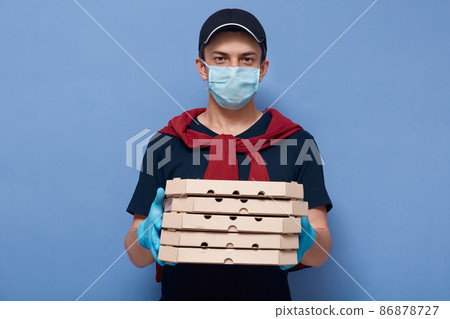 Horizontal indoor picture of charismatic pleasant delivery guy holding ordered food in boxes, wearing gloves and antibacterial mask, having peaceful facial expression. Quarantine time concept. Horizontal indoor picture of charismatic pleasant delivery guy holding ordered food in boxes, wearing gloves and antibacterial mask, having peaceful facial expression. Quarantine time concept. 86878727