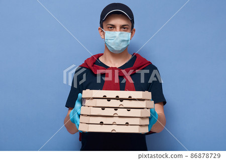 Portrait of serious handsome delivery guy holding carton boxes with food ordered online, working during quarantine, wearing uniform, gloves and protective mask. Coronavirus outbreak concept. Portrait of serious handsome delivery guy holding carton boxes with food ordered online, working during quarantine, wearing uniform, gloves and protective mask. Coronavirus outbreak concept. 86878729