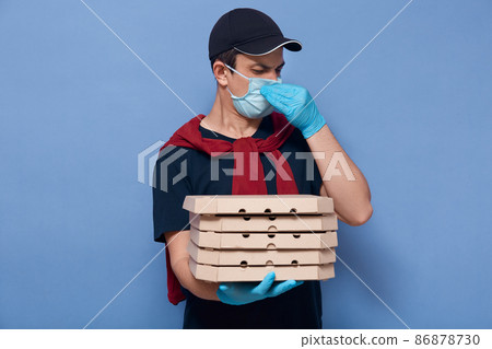 Image of unhappy hard working delivery man holding his nose, having carton boxes in one hand, feeling unpleasant smell from parcel, working during quarantine, wearing mask and gloves. Work concept. Image of unhappy hard working delivery man holding his nose, having carton boxes in one hand, feeling unpleasant smell from parcel, working during quarantine, wearing mask and gloves. Work concept. 86878730