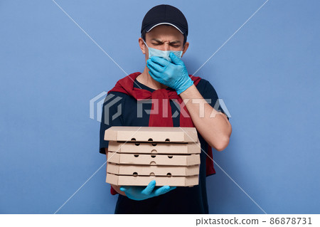 Indoor picture of ill sneezing young courier covering mouth with hand, wearing mask and gloves, closing eyes, being infected with coronavirus, feeling unwell, holding carton boxes in one hand. Indoor picture of ill sneezing young courier covering mouth with hand, wearing mask and gloves, closing eyes, being infected with coronavirus, feeling unwell, holding carton boxes in one hand. 86878731