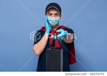 Horizontal shot of astonished male wearing t shirt, sweater over shoulders, cap, protective mask,latex gloves, courier hands black shopping bag to client with shocked expression, keeps palm on cheek Horizontal shot of astonished male wearing t shirt, sweater over shoulders, cap, protective mask,latex gloves, courier hands black shopping bag to client with shocked expression, keeps palm on cheek 86878745